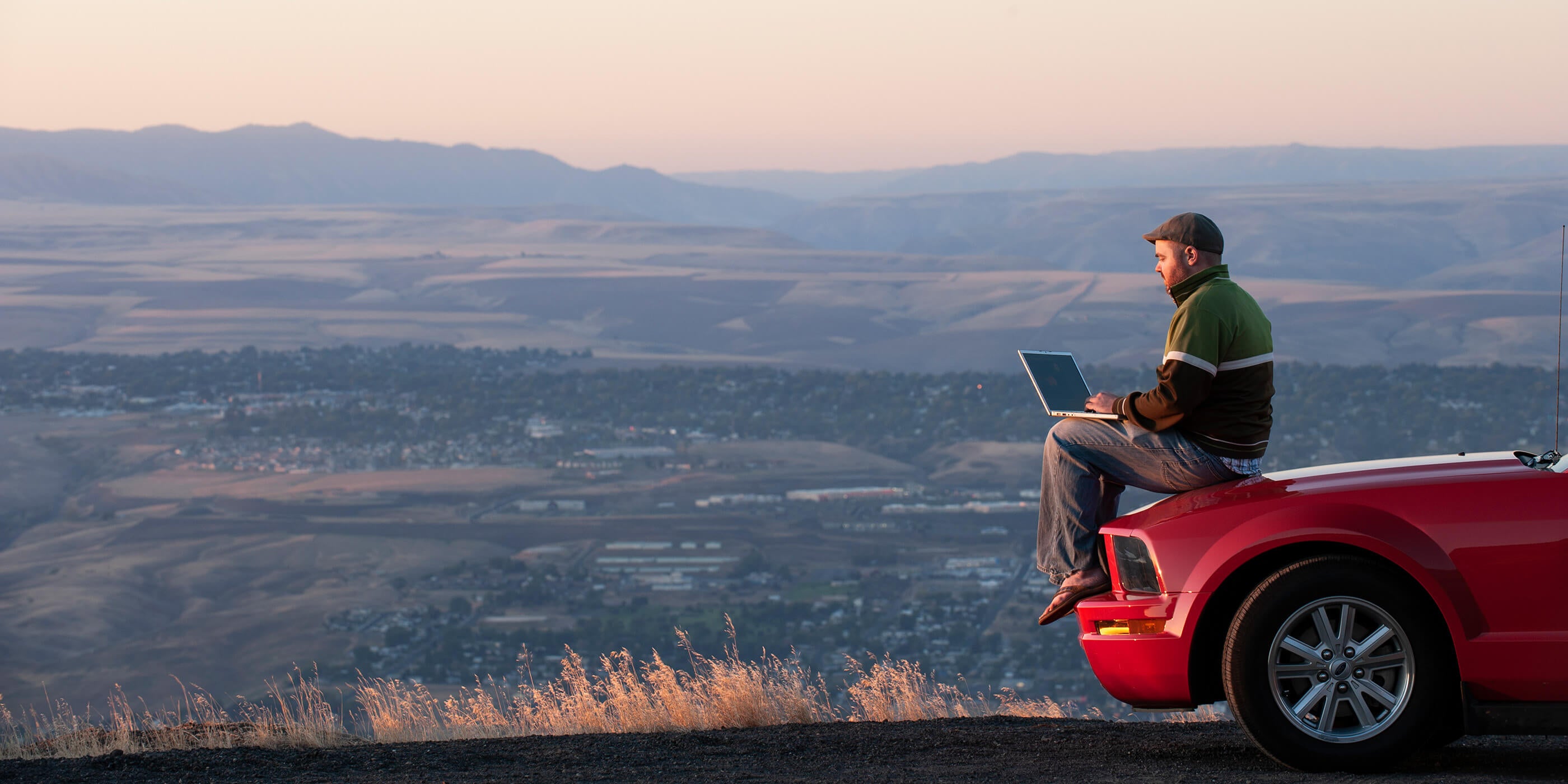 Mann arbeitet am Laptop auf der Motorhaube sitzend vor einer Bergpanorama