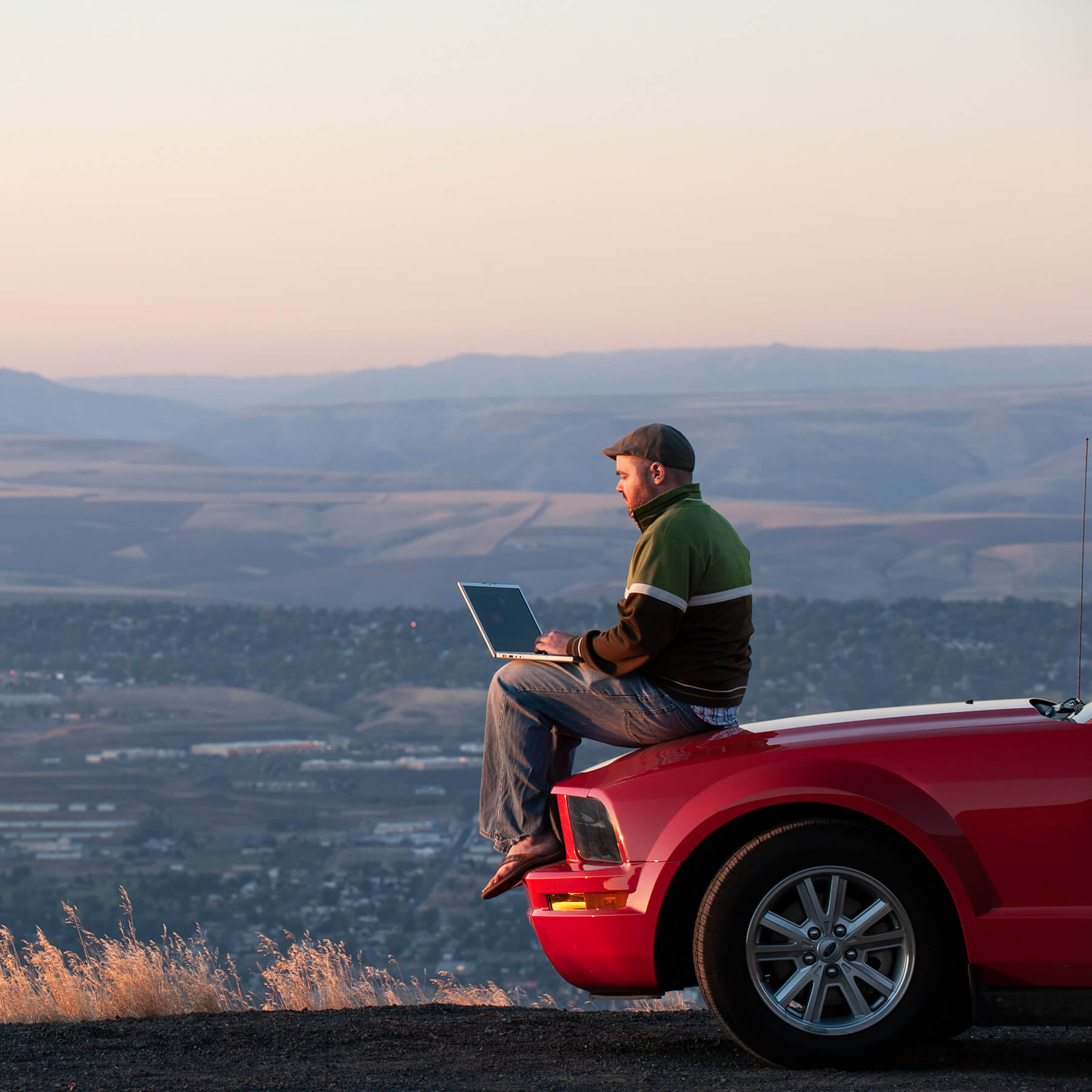 Mann arbeitet am Laptop auf der Motorhaube sitzend vor einer Bergpanorama Mobile Größe